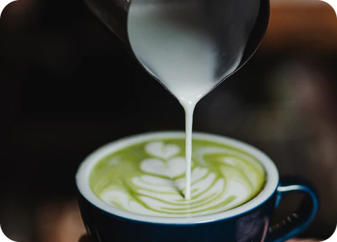 Spoon pouring white liquid into a cup of green matcha latte with decorative patterns.